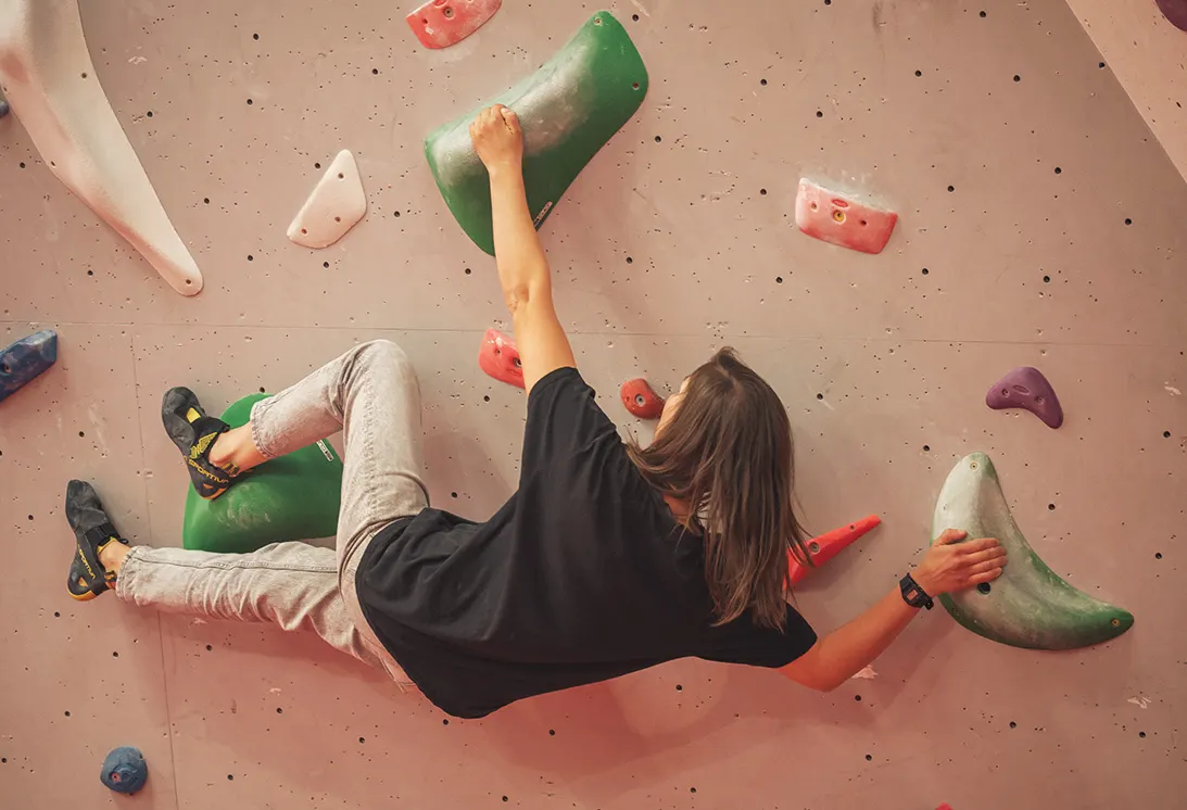 Person climbing an indoor wall with coloured hand and foot holds, viewed from above.