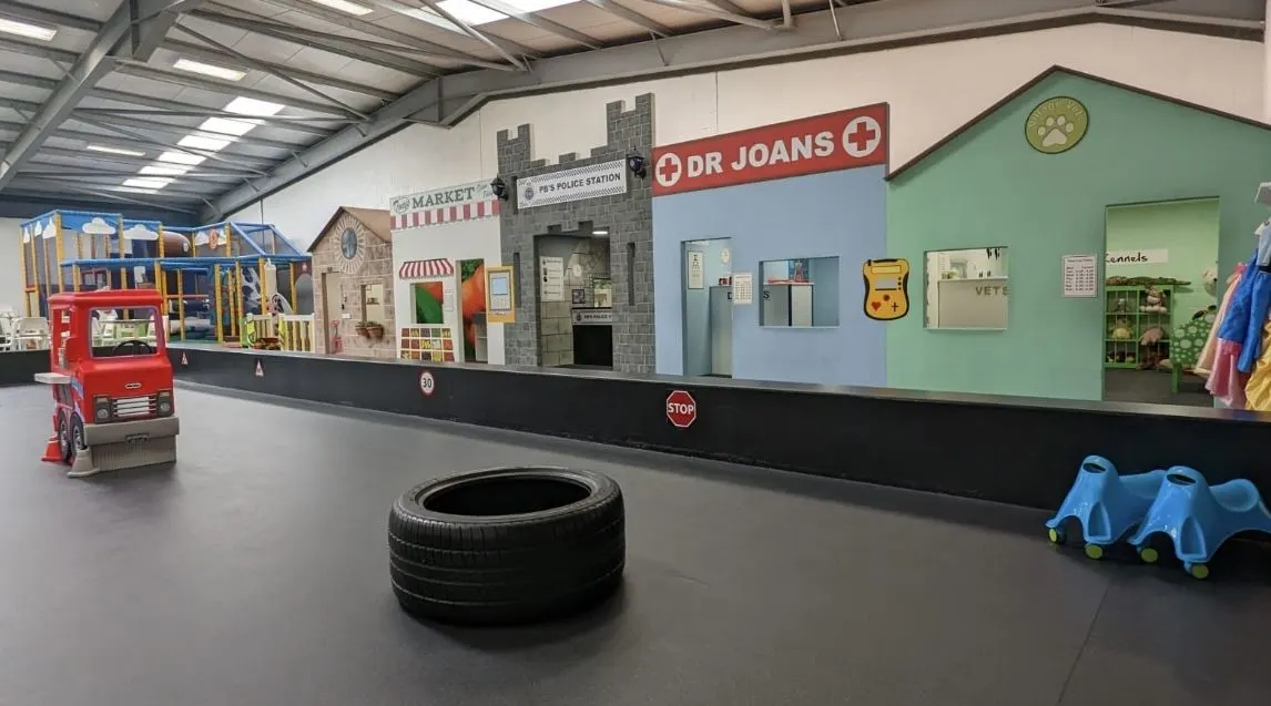 Indoor play area with themed building facades including a market stall, police station and doctor's surgery, with a tyre and blue play equipment on the floor.