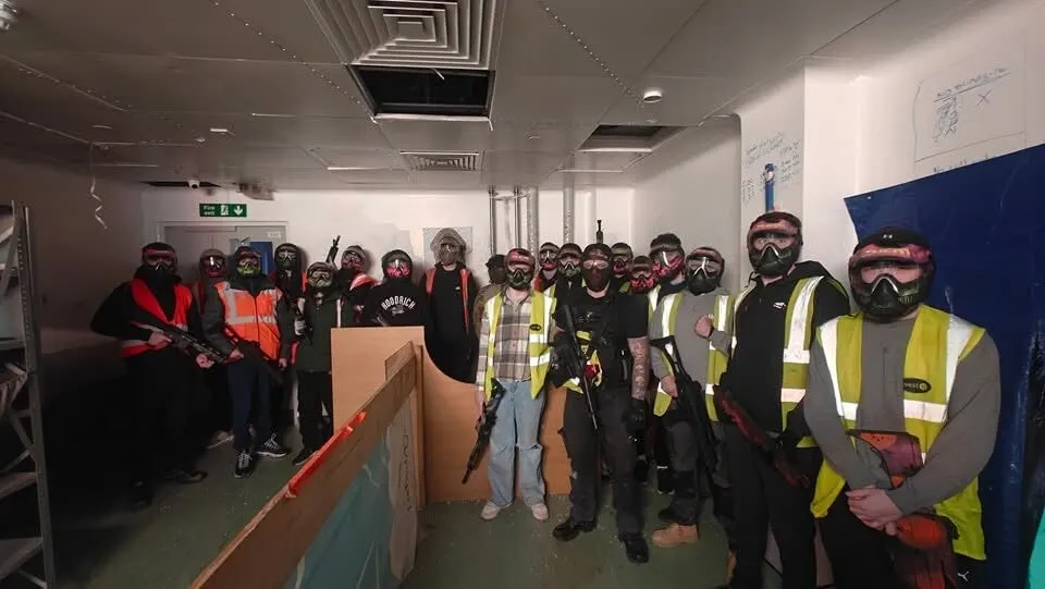Large group of people wearing protective gear and masks posing together in an indoor arena facility.