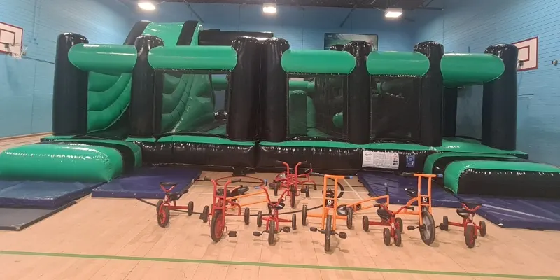 Multiple tricycles arranged on a foam floor in front of green and black inflatable play structures indoors.
