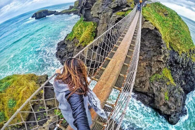 Person crossing a rope bridge suspended between moss-covered cliff faces above turquoise sea.