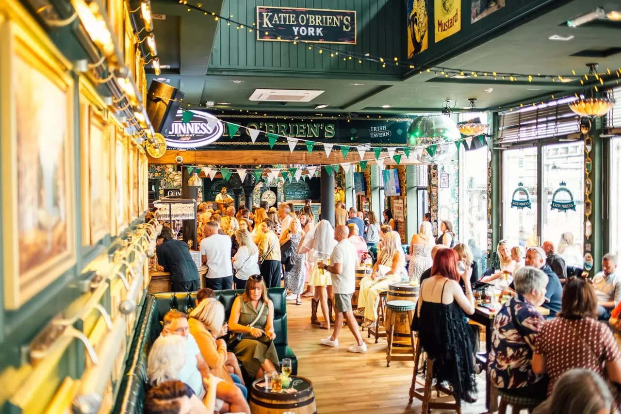 Busy Irish pub interior with crowds of people standing and seated at tables, decorated with green and gold bunting and beer brand signage.