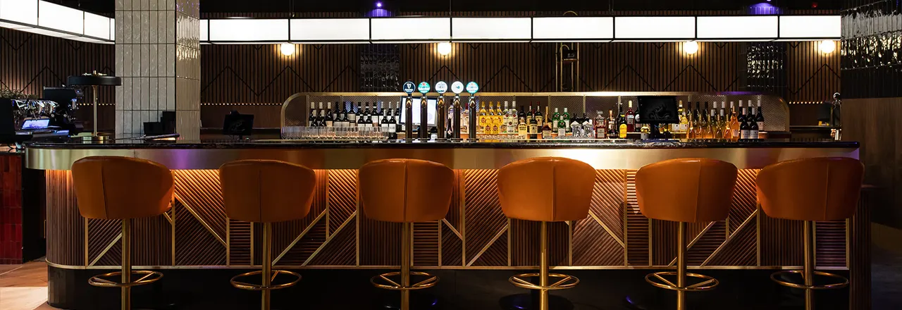 Modern bar counter with empty orange velvet bar stools and illuminated shelving displaying spirits and cocktail glasses.