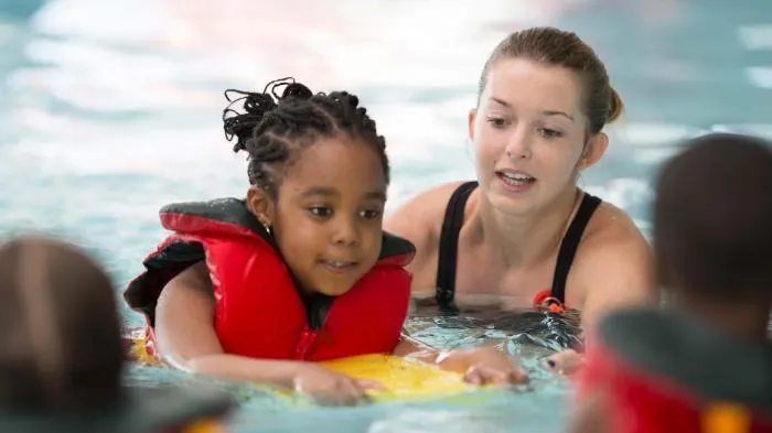 A young child wearing a red buoyancy aid learns to swim with an instructor in a pool.
