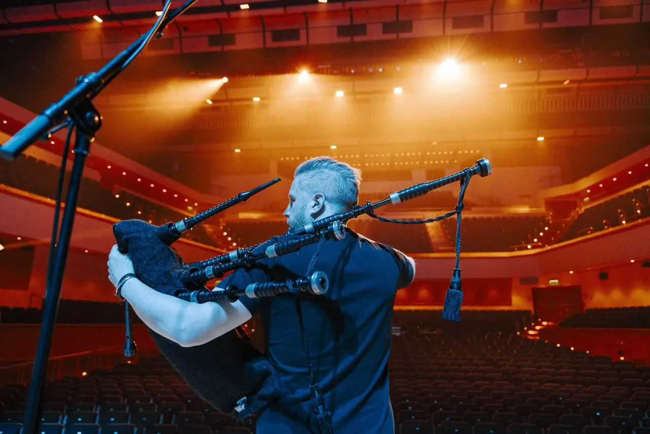 Musician playing bagpipes on stage in a concert hall with warm stage lighting.