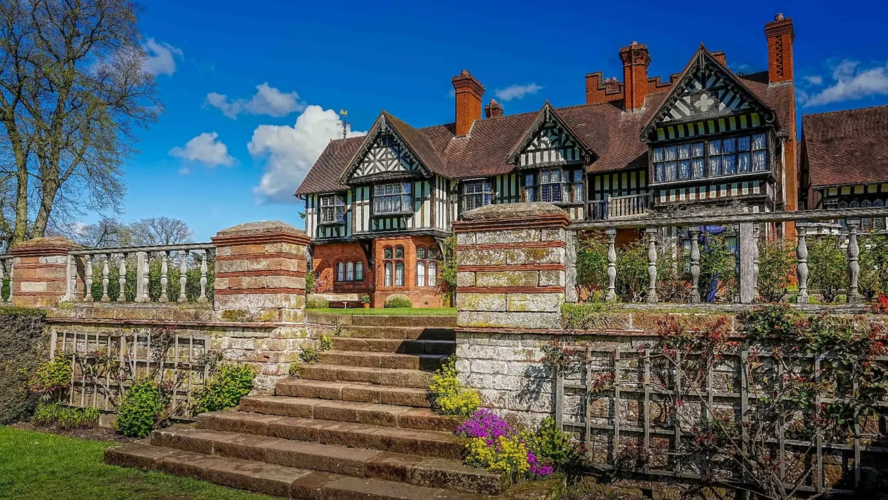 Historic Tudor-style manor house with black and white timber framing, viewed from formal gardens with stone balustrades and flowering shrubs.