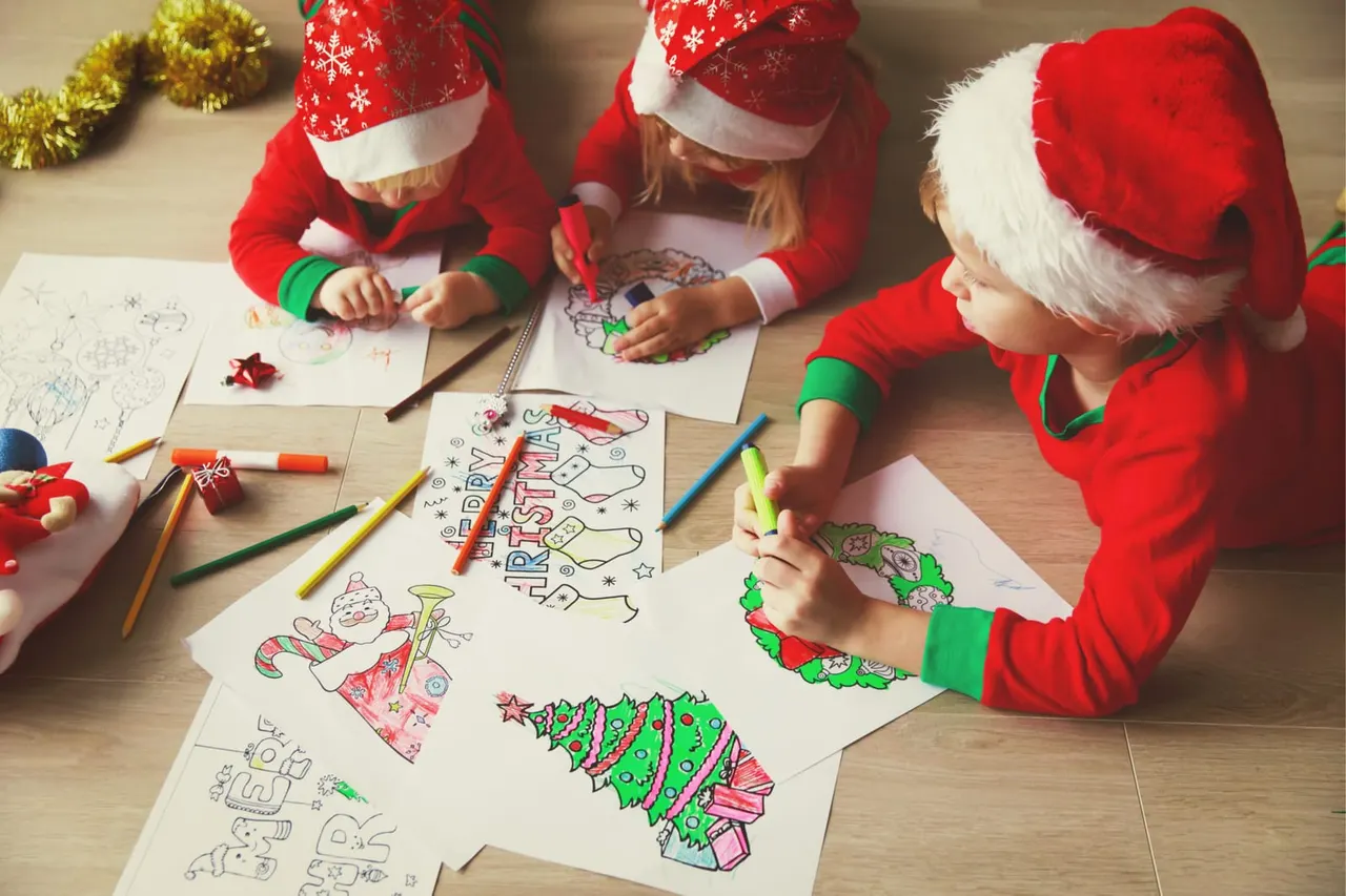 Three children wearing Christmas jumpers and Santa hats colouring festive drawings with markers at a wooden table.