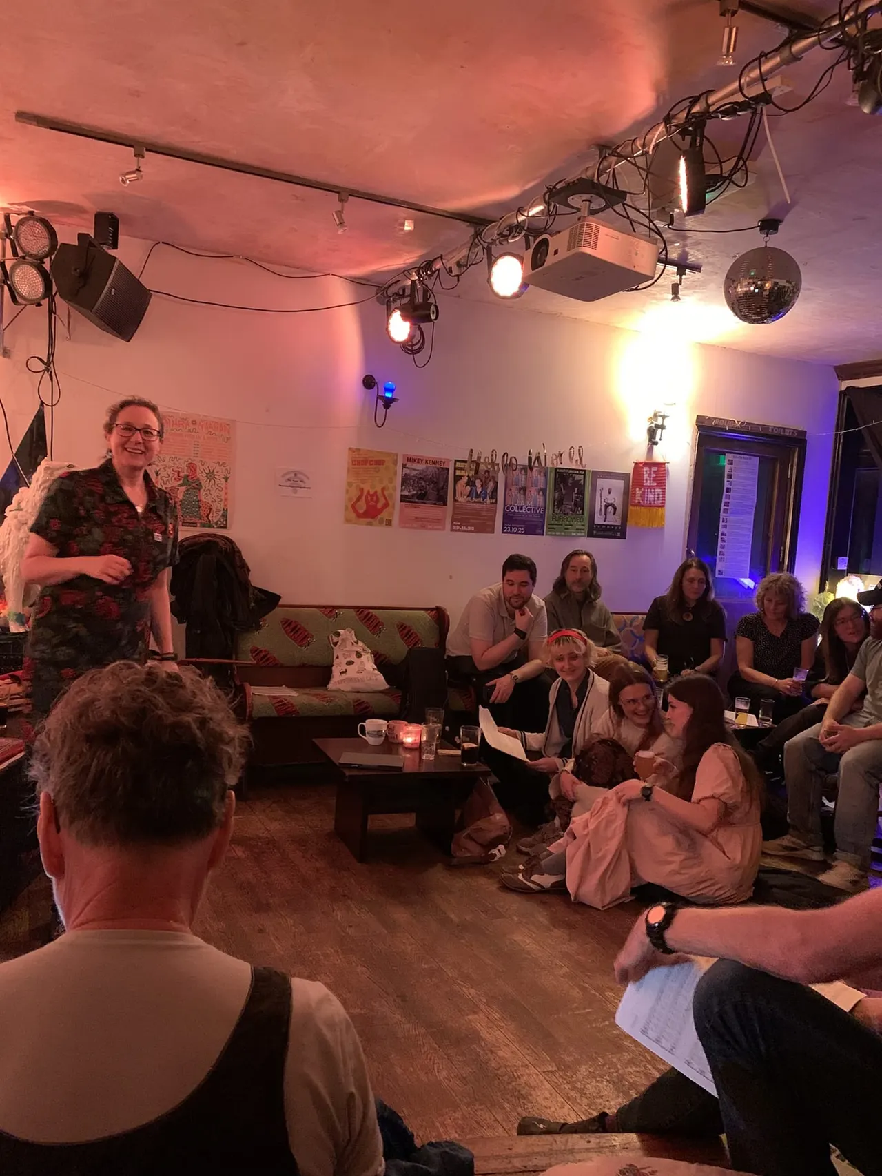 A group of people gathered in a venue with purple and pink lighting, seated around a sofa and floor whilst one person stands presenting.