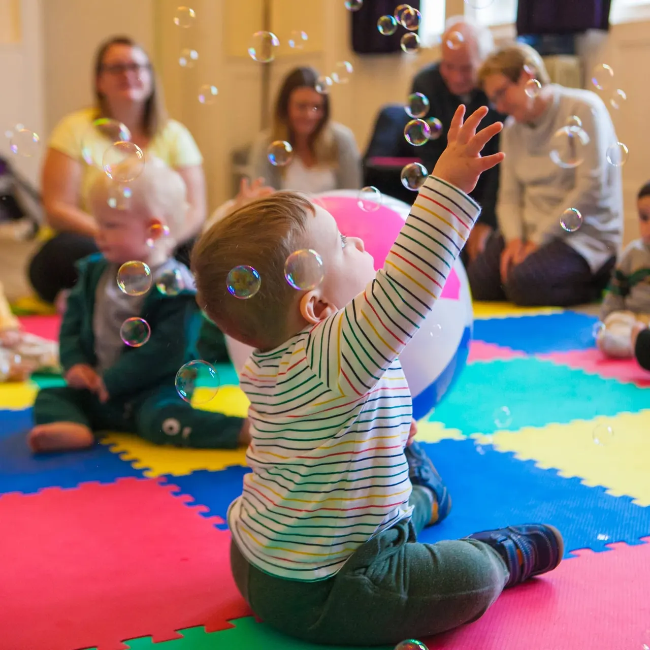A toddler in a striped top sits on a colourful play mat, reaching up at bubbles whilst adults and children watch nearby.