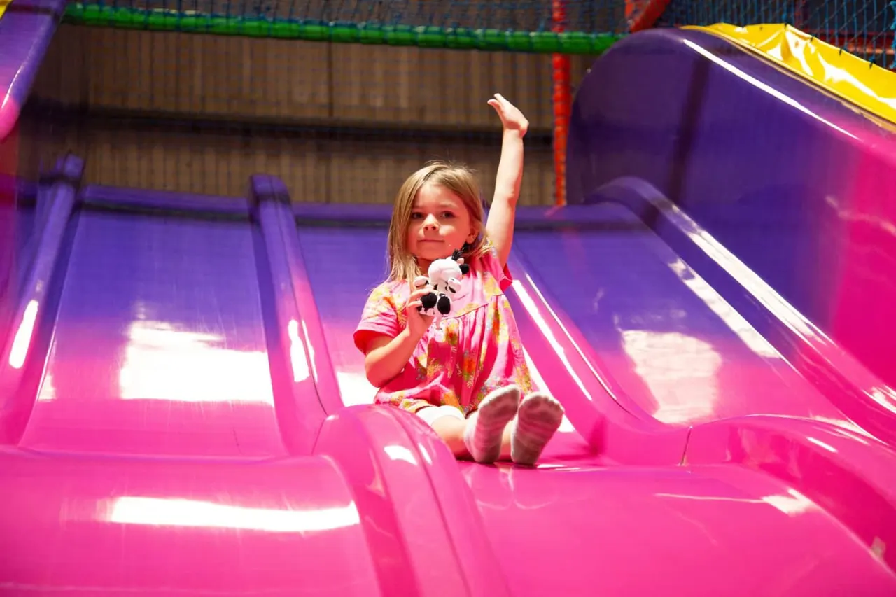 Young child sitting inside a brightly lit pink inflatable play structure, holding a stuffed toy and waving.