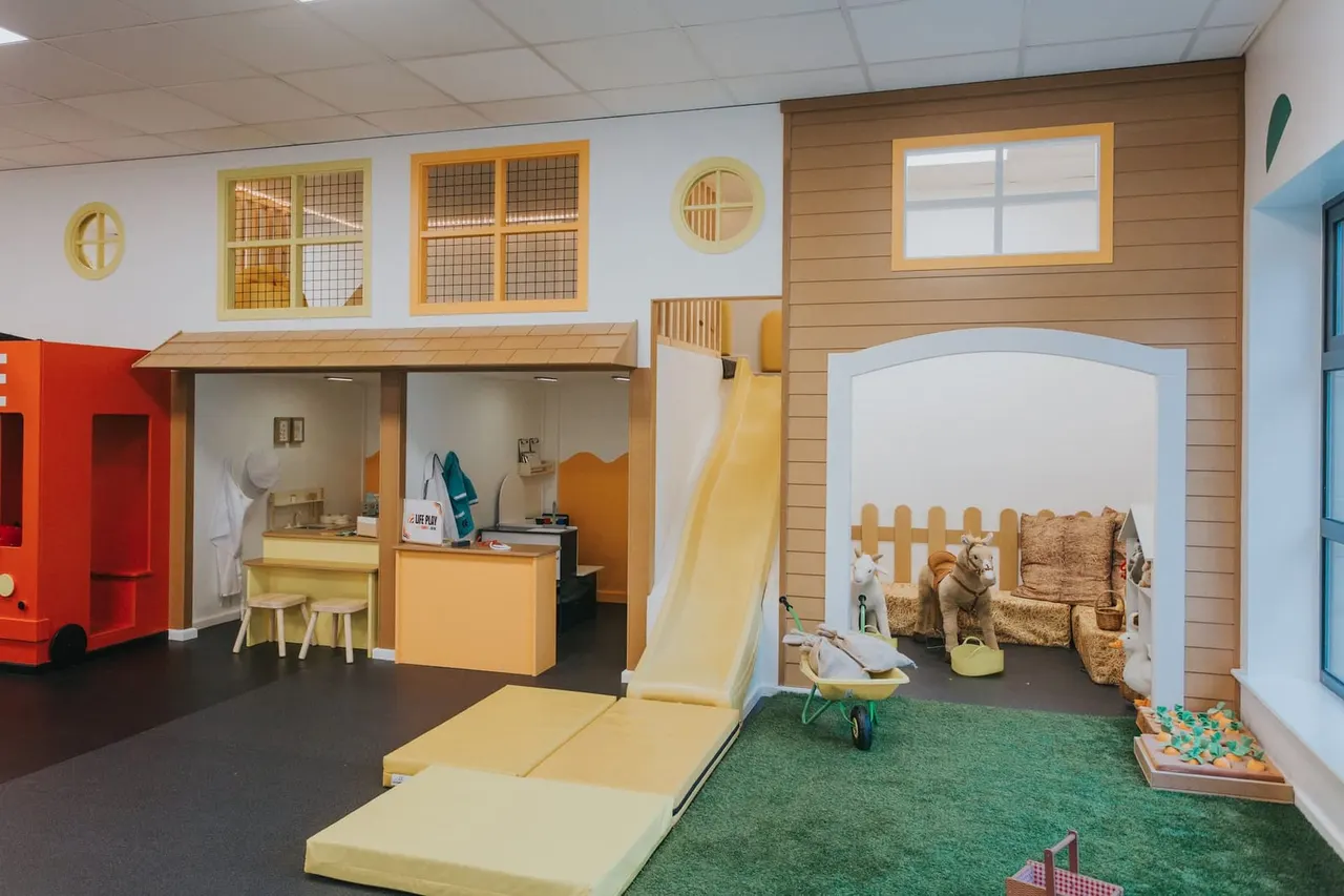 Indoor play area with wooden play structures including a slide, kitchen unit, and cottage-style room with seating and toys for young children.
