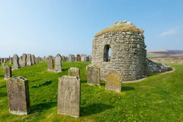 Historic stone cemetery building surrounded by weathered gravestones on grassy hillside under blue sky.