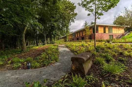 A winding path through wooded grounds leads to a yellow building surrounded by trees and landscaping.