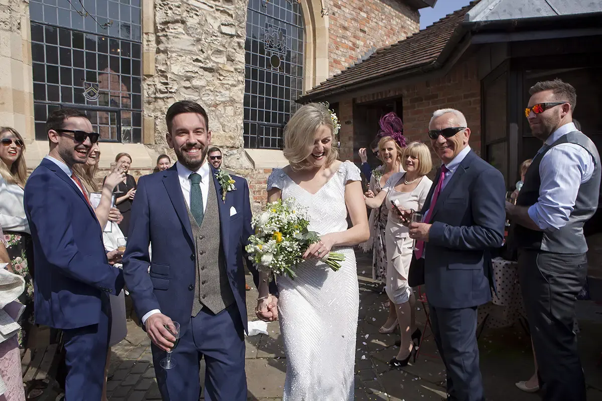 Bride and groom walk together holding hands whilst smiling at wedding guests outside a stone building.