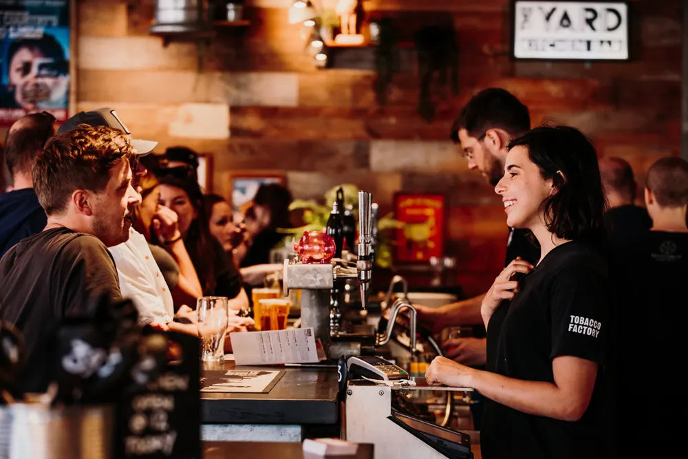 Bartender serving drinks to customers at a wooden bar counter inside a warm-lit cafe venue.