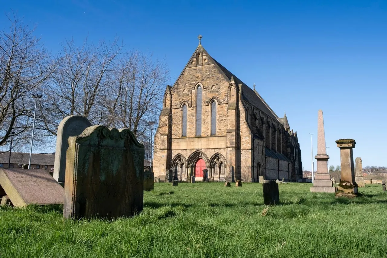 Historic stone church with graveyard, weathered headstones and monuments scattered across grass under clear blue sky.