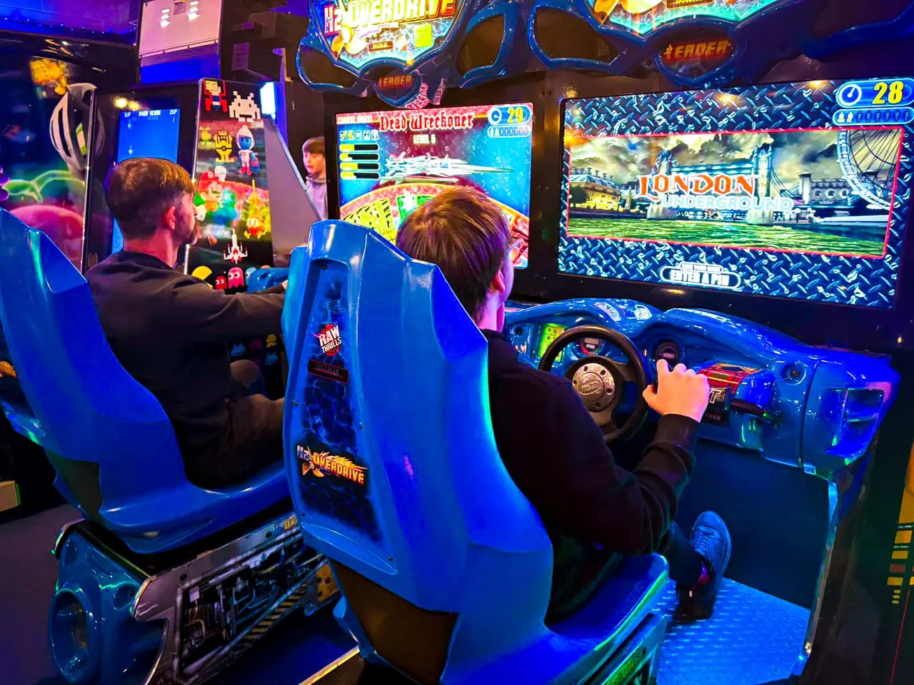 Two players seated in a bright blue racing arcade machine surrounded by other colourful arcade games in a dimly lit gaming venue.