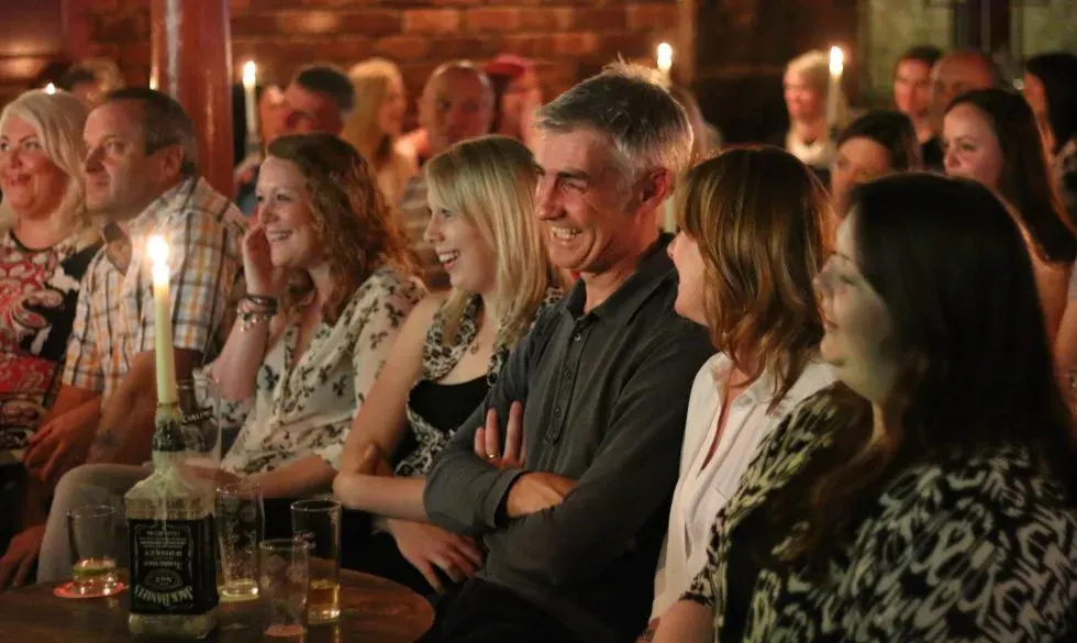 A diverse group of adults sitting at a bar counter, smiling and watching a performance in a warmly lit brick venue.