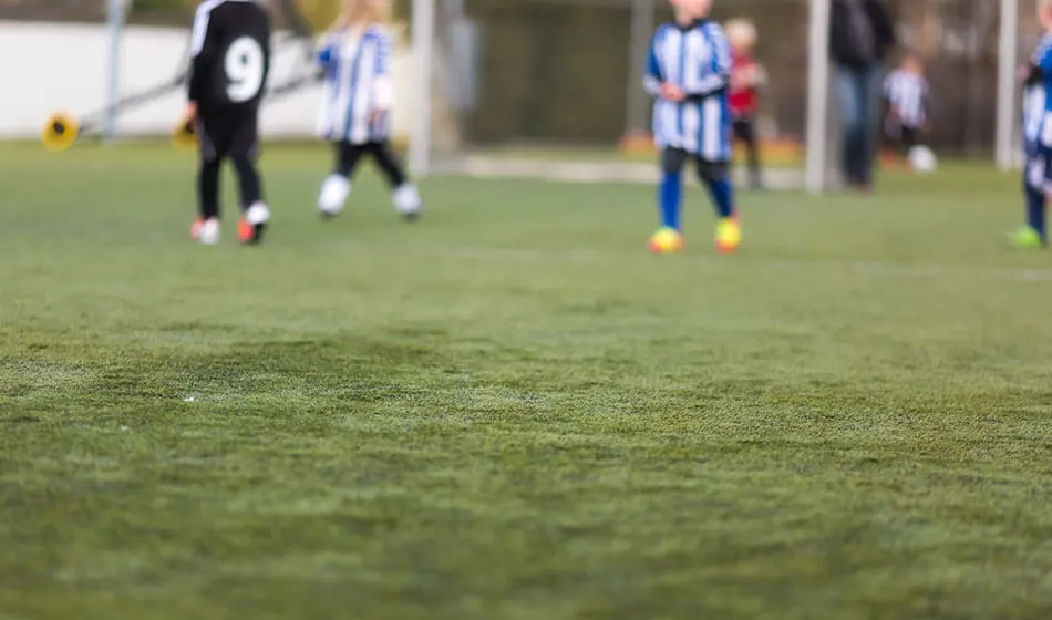 Children playing football on a grass pitch with training cones and spectators watching from the sidelines.