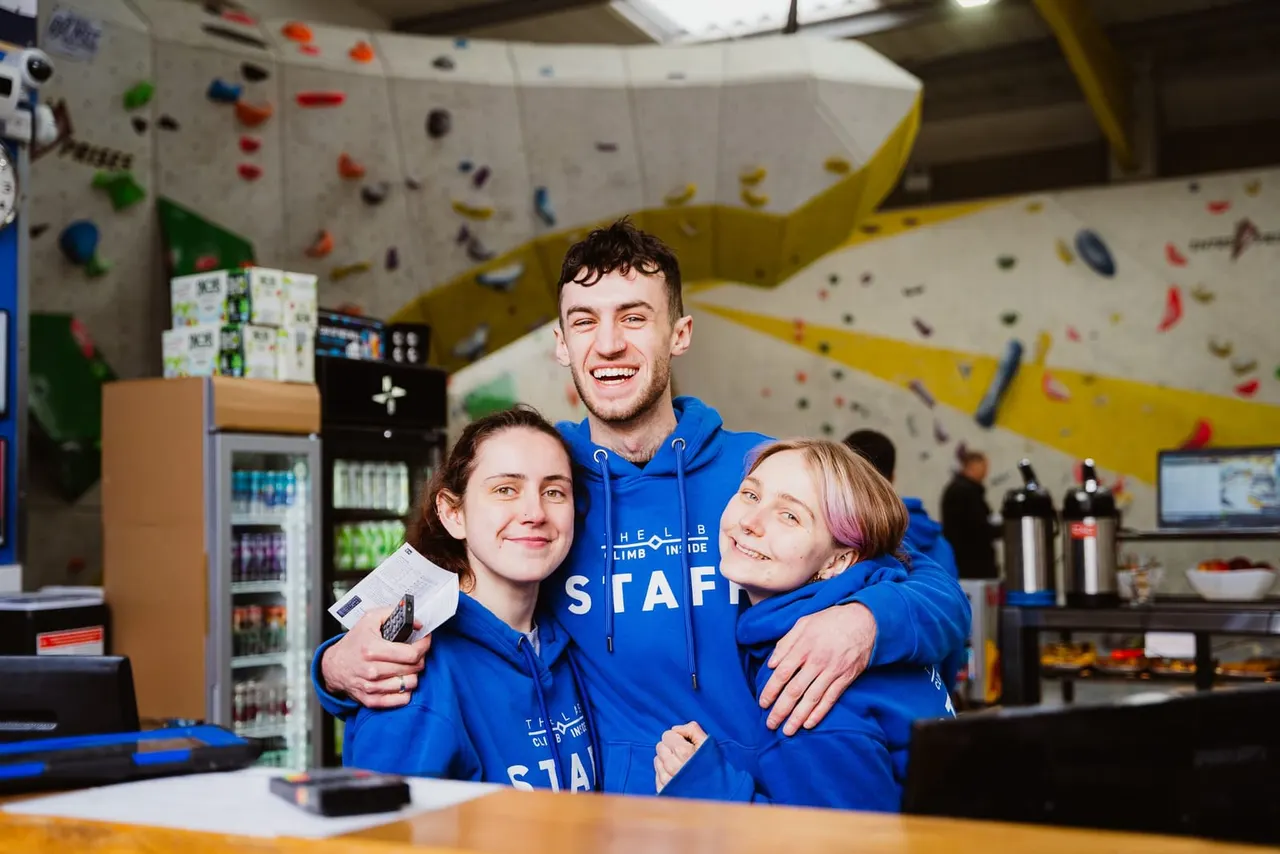 Three smiling staff members in blue hoodies pose together inside an indoor climbing wall facility with colorful holds.