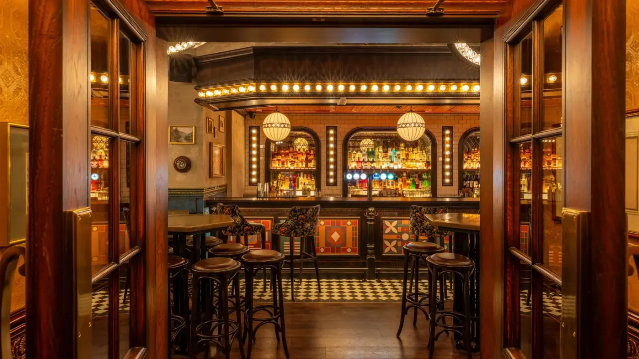 A vintage-style bar interior with high tables and stools facing an ornate backlit bar with shelves of spirits and decorative globe pendant lights.