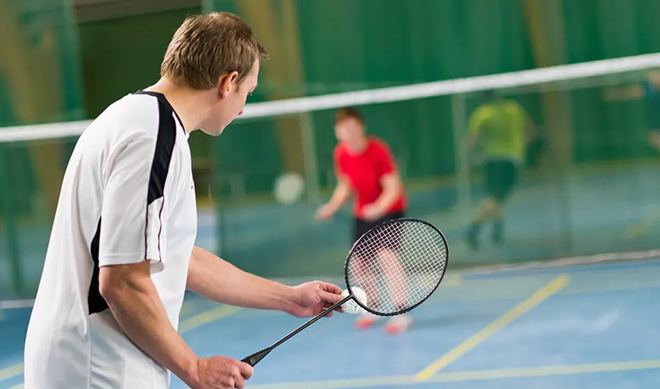 Man in white shirt holding a tennis racket on an indoor court with opponent in red in the background.