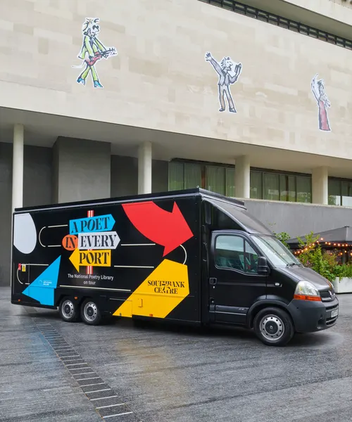 A black mobile library van with colourful geometric designs parked outside a modern building with illustrated figures on its facade.