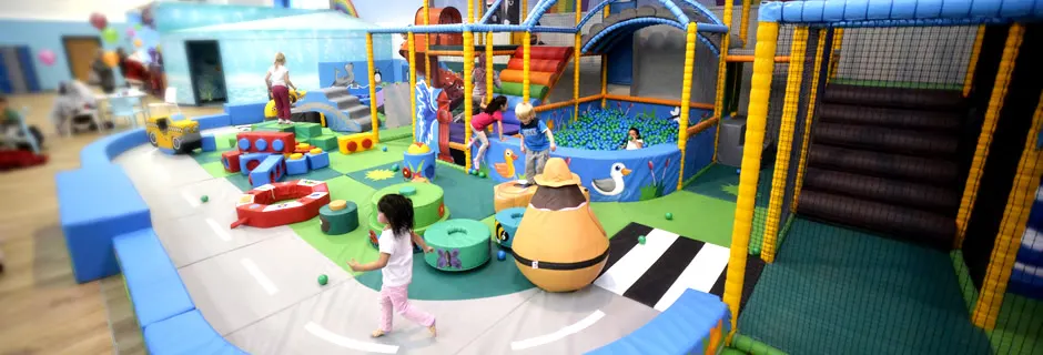 Young children playing in a colourful indoor play area with soft structures, ball pits and climbing equipment.