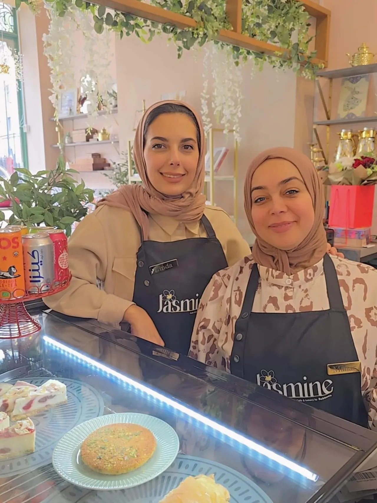 Two staff members standing behind a counter at a café, with pastries on display and hanging greenery decoration.