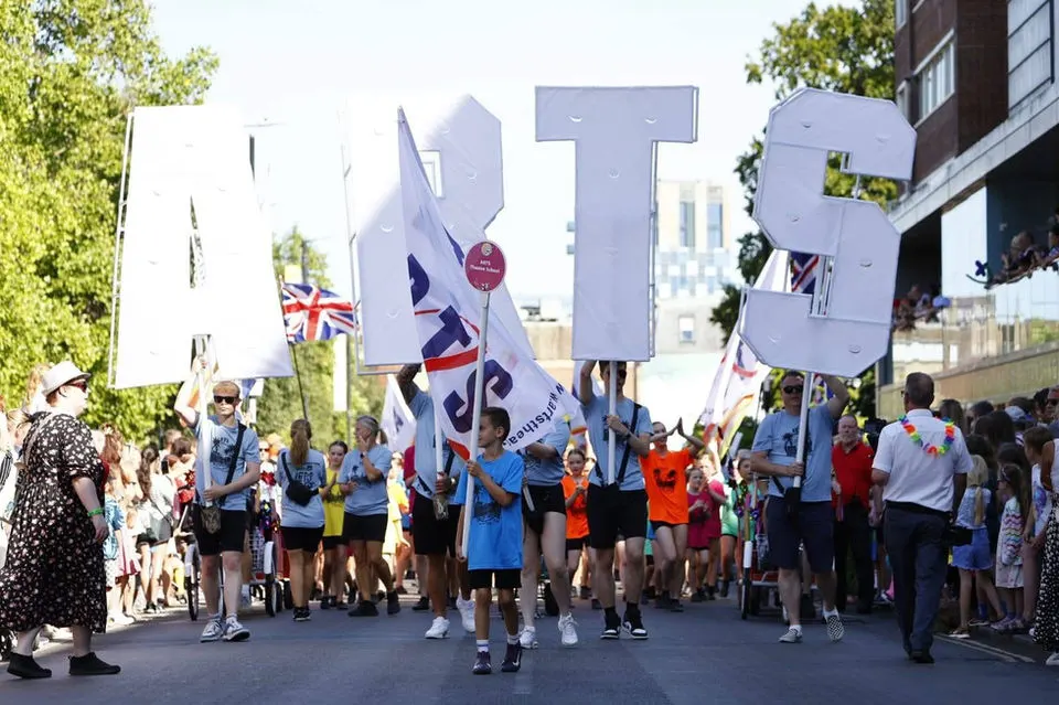 A large group of children and young people parading through a street carrying oversized letters and flags during a celebratory event.