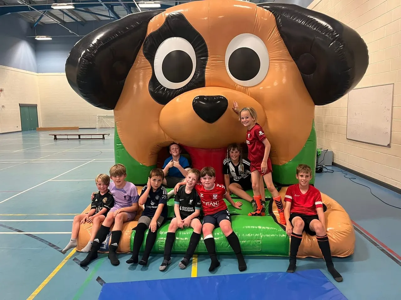 Children posing in front of a large inflatable dog mascot in an indoor sports hall.