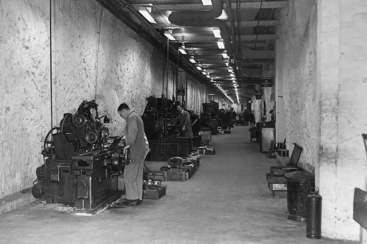 Workers operating industrial machinery in a long, concrete underground tunnel with overhead lighting.