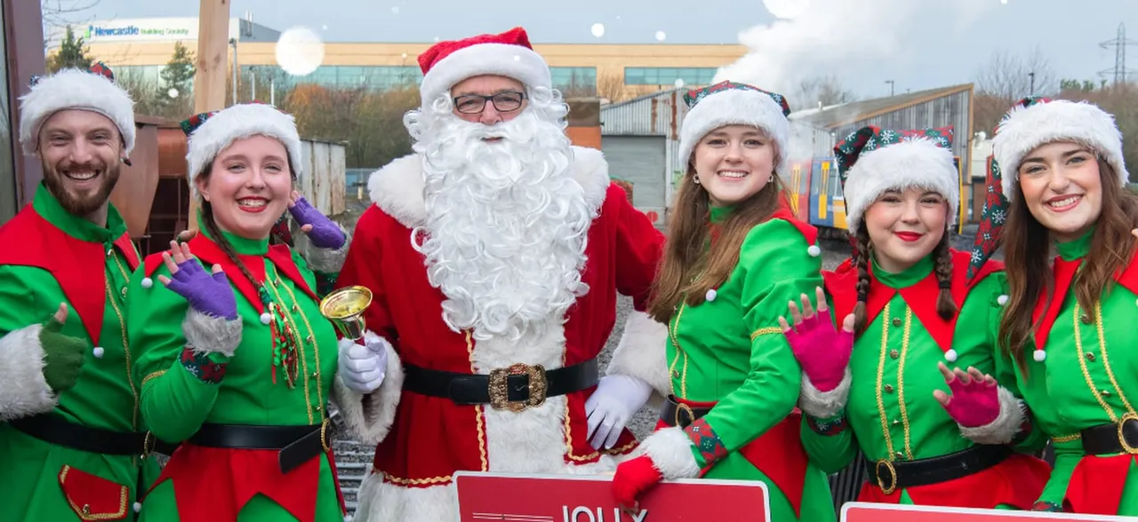 Father Christmas and six elves in festive costumes pose together outdoors at a Christmas event.