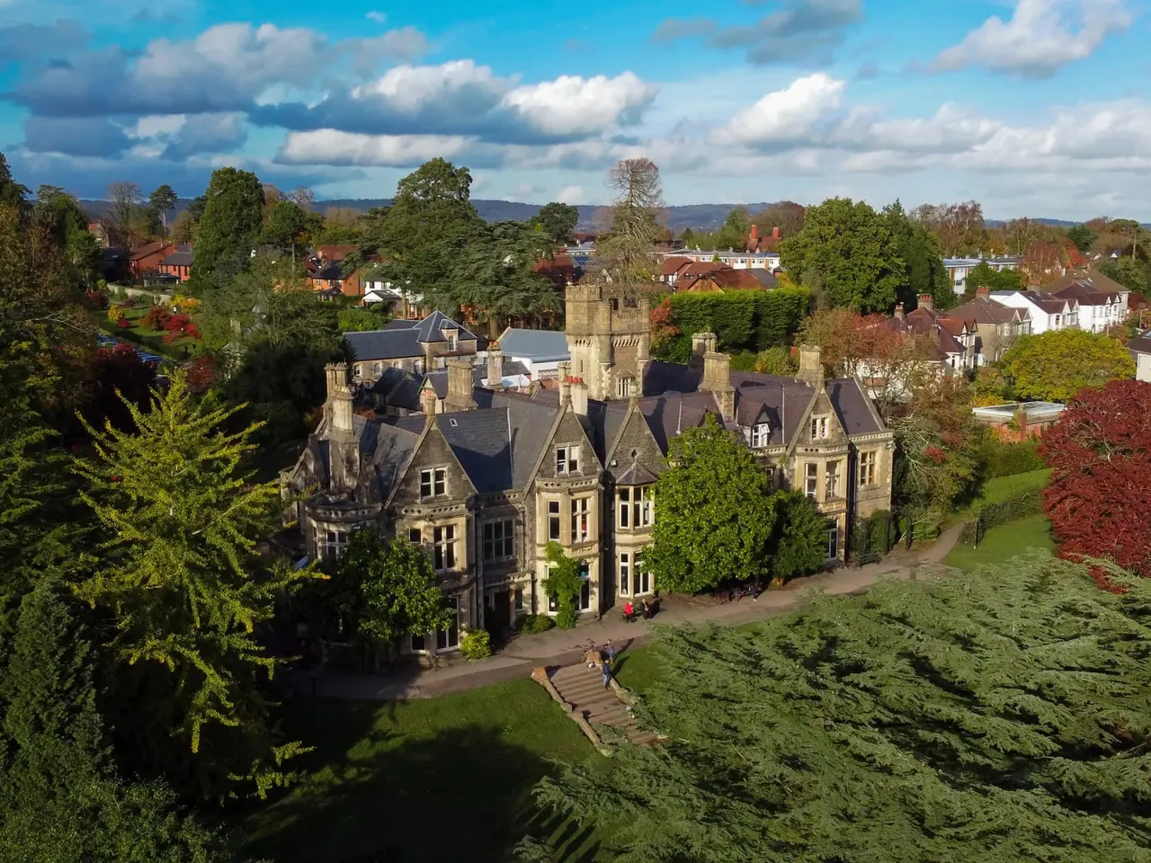 Victorian mansion with Gothic towers set in extensive grounds, surrounded by trees and countryside with hills in the distance.