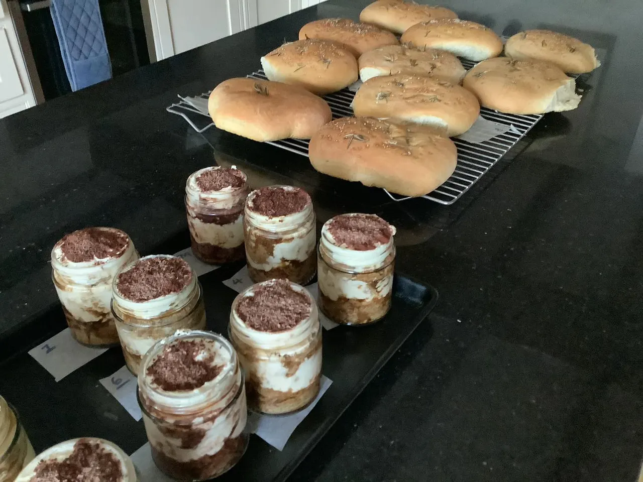 Freshly baked bread rolls with herbs on a cooling rack and tiramisu desserts in jars displayed on a dark counter.