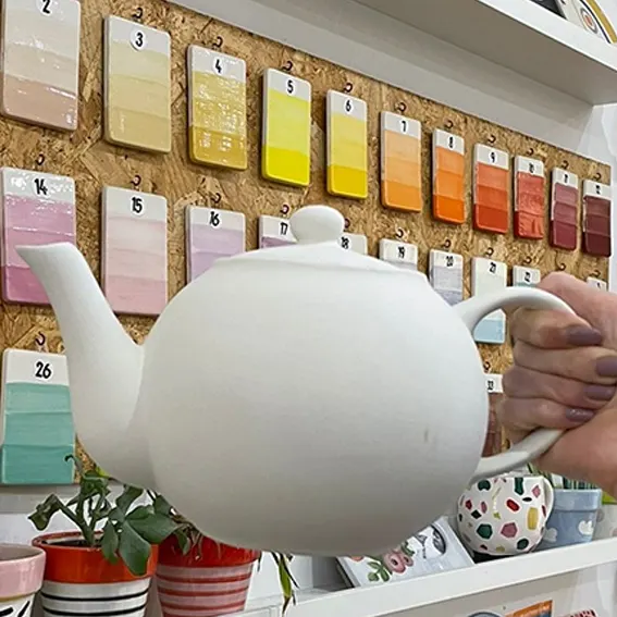 Person holding a large white ceramic teapot in front of a cork board displaying numbered paint colour swatches.