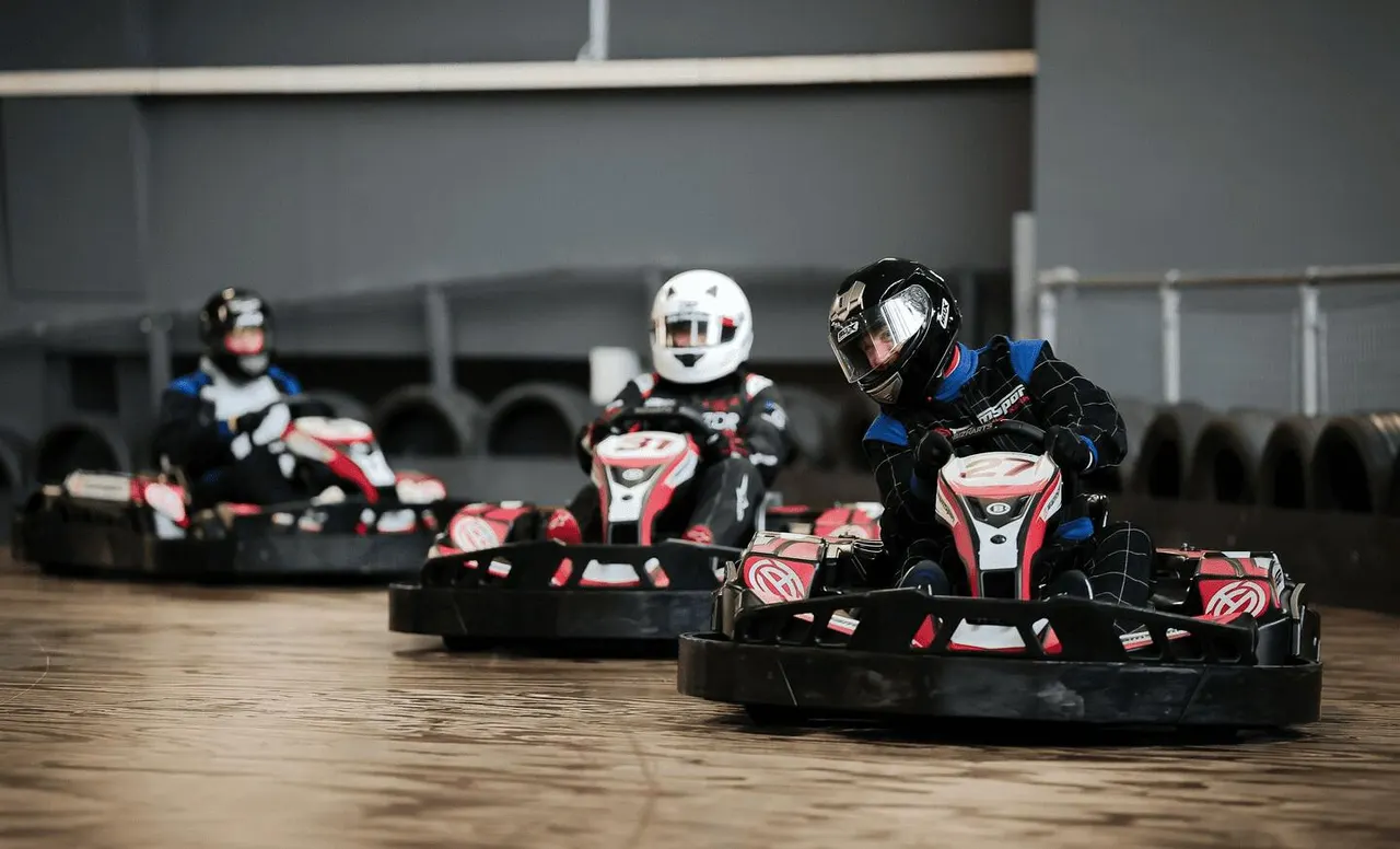 Indoor go-kart race with multiple drivers in helmets competing on a track inside a warehouse facility.