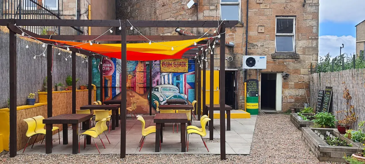 Colourful outdoor courtyard seating area with yellow and brown chairs beneath a pergola with red and yellow fabric canopy, decorated with hanging lights.