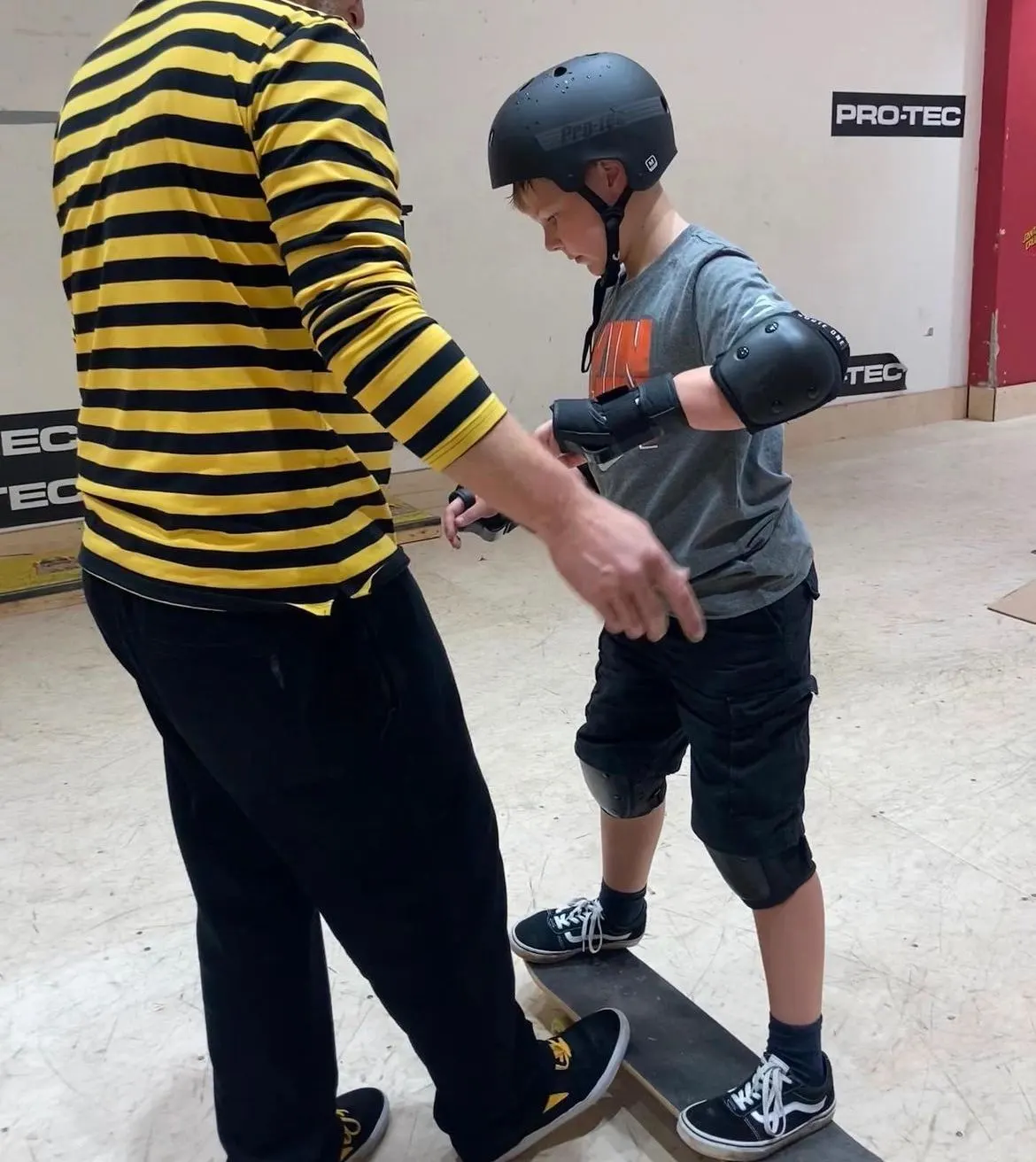 Young person wearing protective gear learning to skateboard with an instructor in striped shirt indoors.