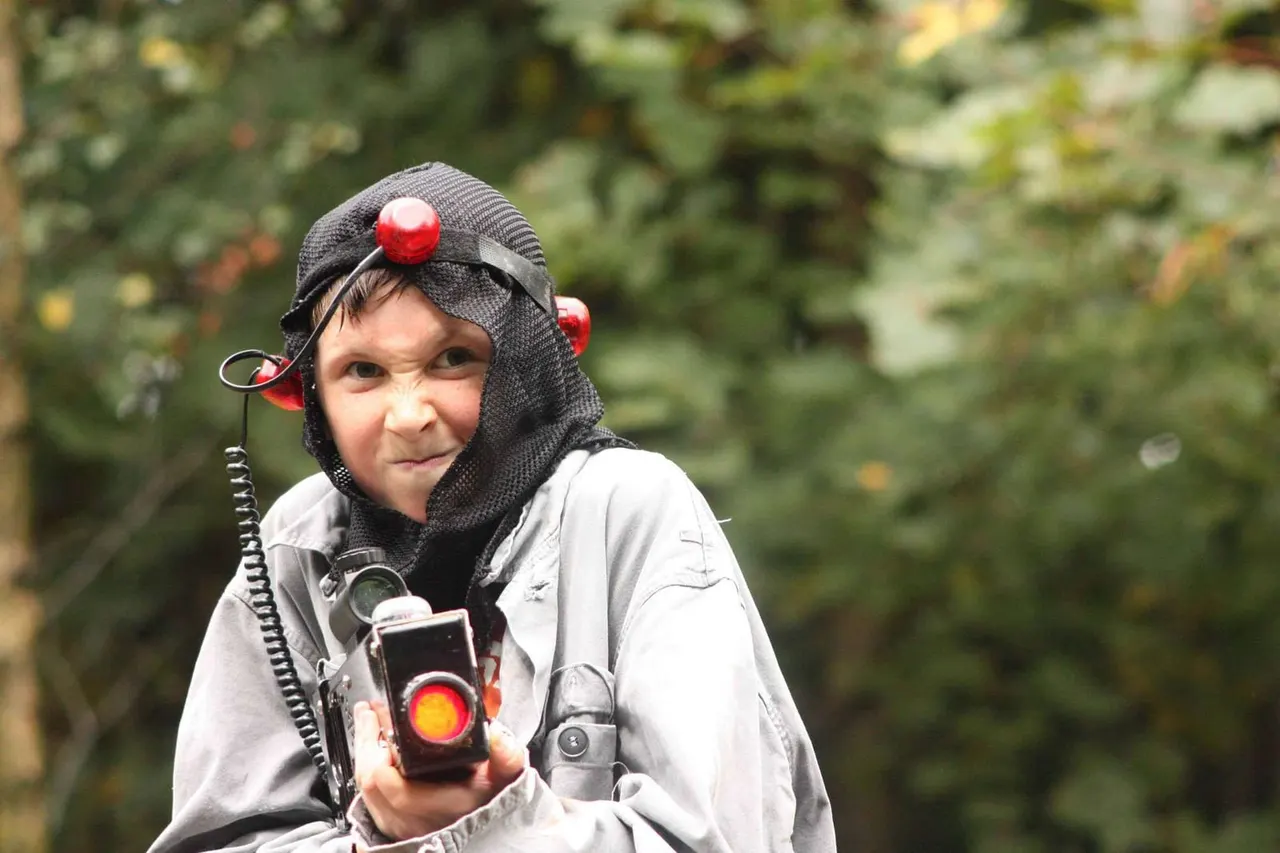 Child in paintball gear holding a marker gun, wearing a black hood with red pompoms, against a foliage background.