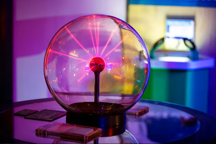 Plasma ball with pink and purple filaments glowing inside a clear glass sphere on a dark base, surrounded by coloured lighting.
