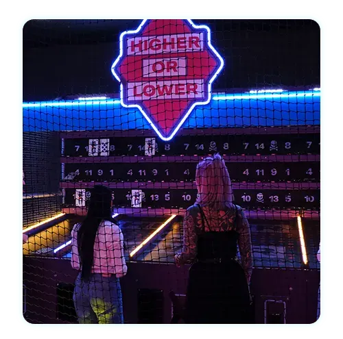 Two people stand facing a neon-lit arcade game installation with a large "higher or lower" sign glowing above.