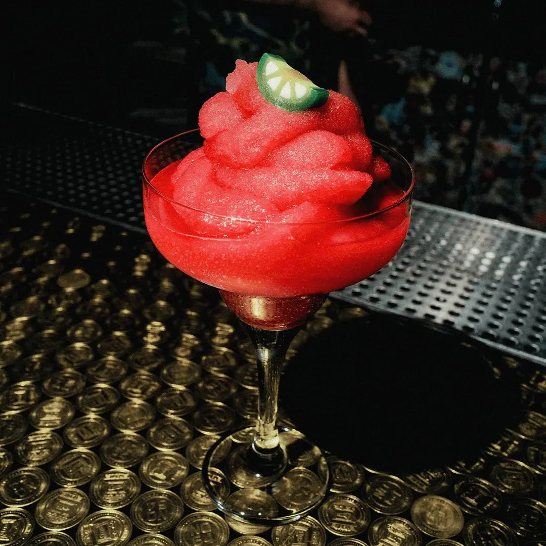 A bright red frozen cocktail topped with crushed ice and a lime slice, served in a coupe glass on a bar counter.