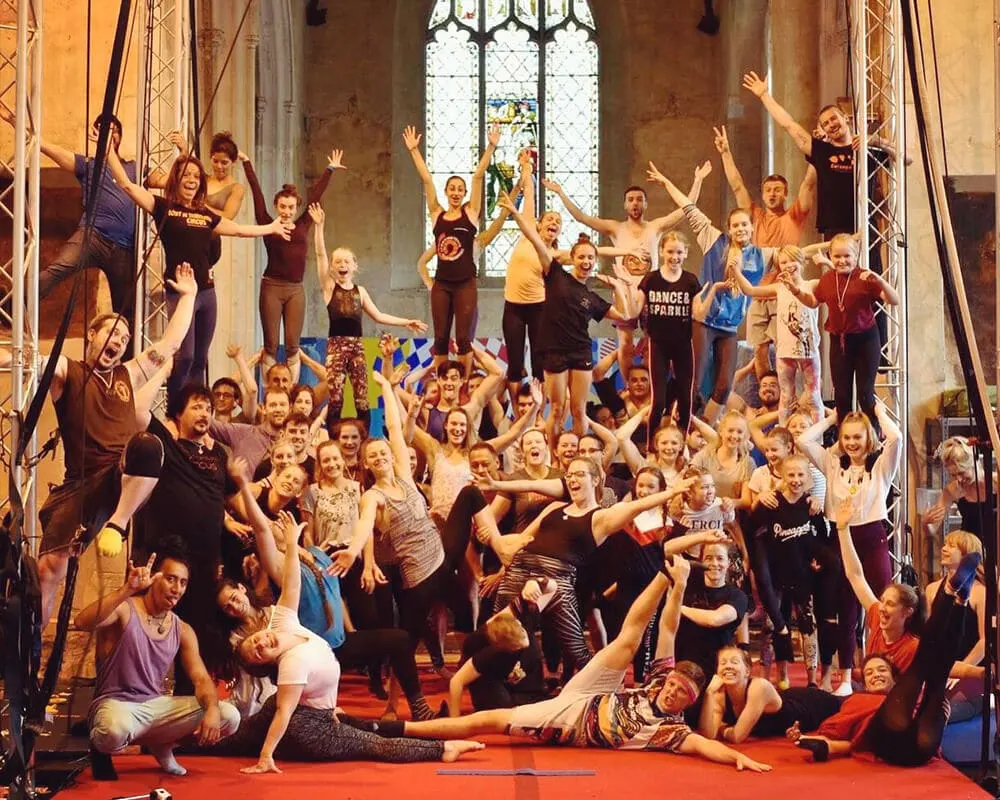 Large group of children and young people dancing and posing with raised arms inside a historic stone hall with tall stained-glass windows.