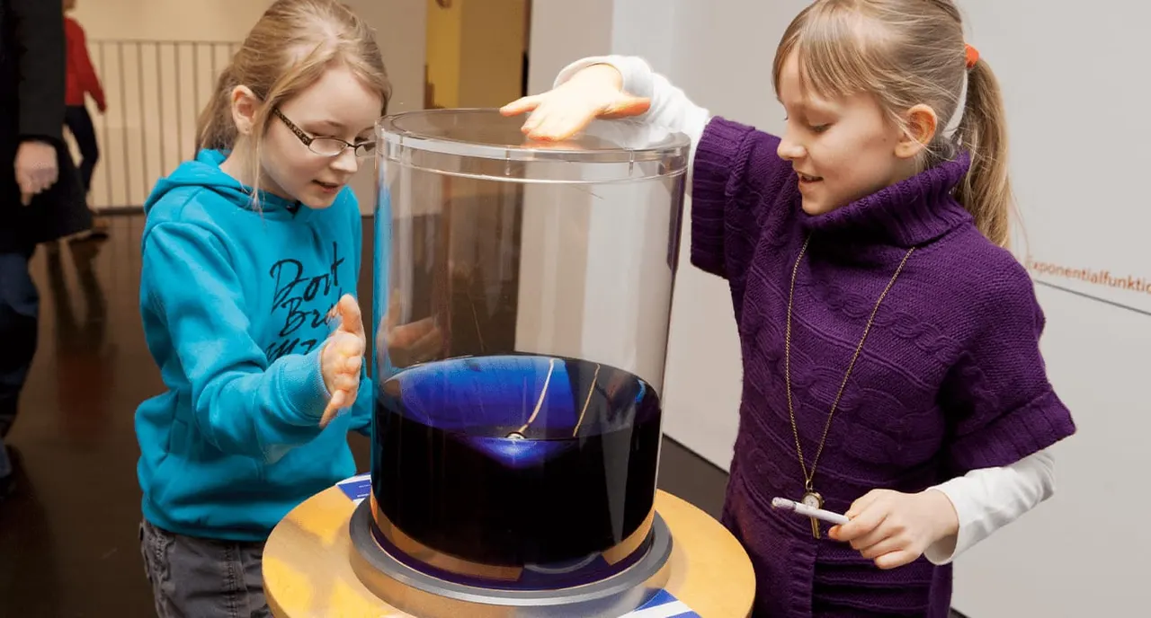 Two girls interact with a large interactive science exhibit containing dark blue liquid and illuminated elements indoors.