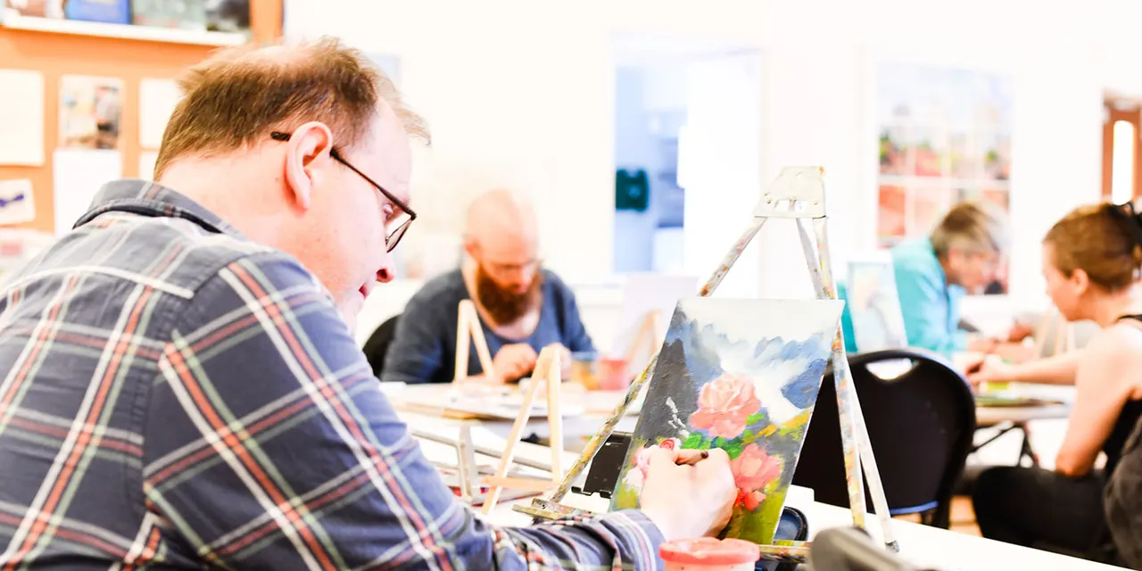 A young person wearing glasses and a checked shirt paints at an easel during an art class with other participants in the background.