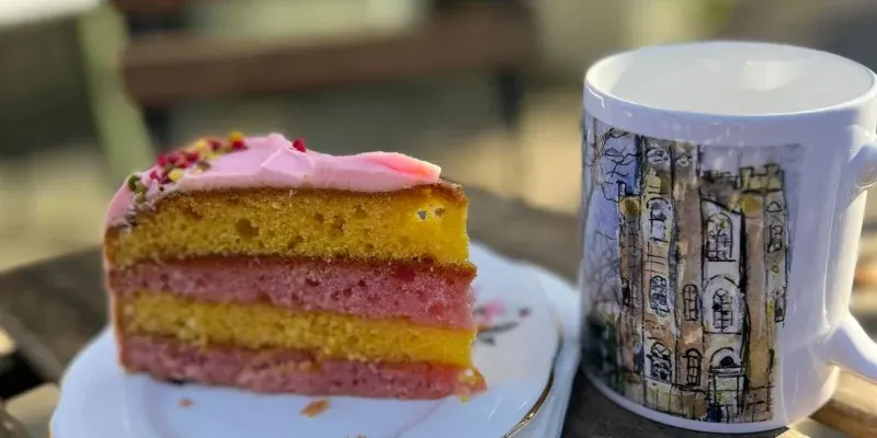A slice of layered sponge cake with pink frosting and sprinkles on a plate beside a decorative mug outdoors.