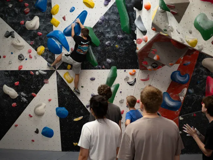 Person climbing an indoor wall with coloured holds whilst others watch from below.