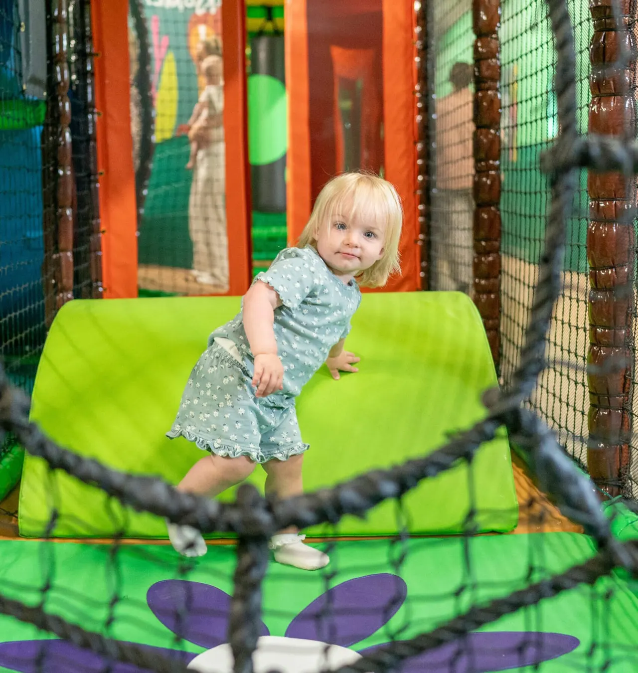A young toddler with blonde hair playing on a bright green bouncy structure inside an indoor soft play centre.