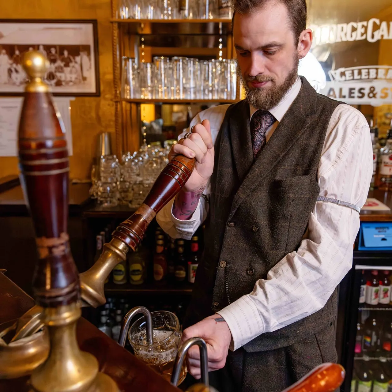 Bartender in waistcoat and tie pouring a drink behind a bar with shelves of glasses and bottles.